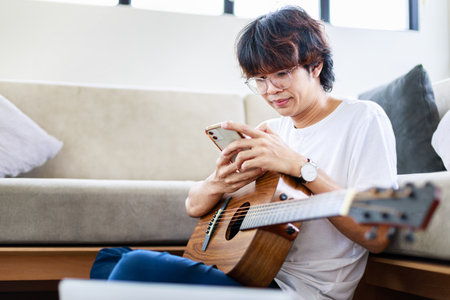 Young man use smartphone while play guitar while sitting on sofa in light living roomの写真素材