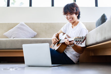 Young man playing acoustic instrumental guitar and sitting on floor in living home at homeの写真素材