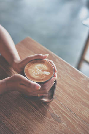 Woman hand hold cup of coffee latte on the wood texture   in vintage color toneの写真素材