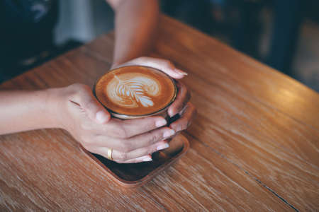Woman hand hold cup of coffee latte on the wood texture   in vintage color toneの写真素材