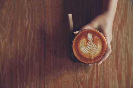 Woman hand hold cup of coffee latte on the wood texture   in vintage color toneの写真素材