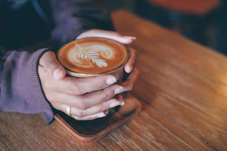 Woman hand hold cup of coffee latte on the wood texture   in vintage color toneの写真素材