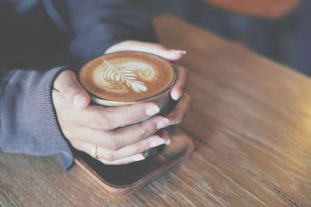 Woman hand hold cup of coffee latte on the wood texture   in vintage color toneの写真素材