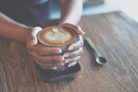 Woman hand hold cup of coffee latte on the wood texture   in vintage color toneの写真素材