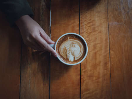 coffee latte art in coffee shop wood texture background with woman hand in vintage color toneの写真素材