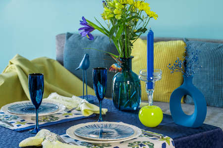 Table setting for two in blue and yellow colors against the background of a sofa with pillows.