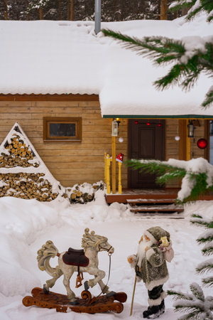 Toy Santa Claus with a wooden rocking horse in the snow in the yard of a wooden houseの写真素材