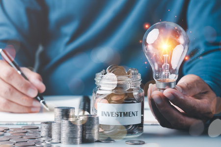 light bulb in a businessman's hand and a pile of coins and coins in a glass jar.All on table,money saving and investment ideas and profit growth,financial management and business strategy.for successの写真素材
