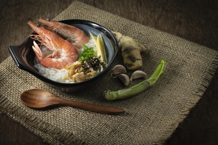 Rice gruel mixed with prawn and ginger, scallion slice and deep fried garlic and black pepper in black bowl. spices put right side and wood spoon put in front of on Sack and wood backgroundの写真素材