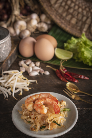 Thai fried noodles or pad thai with shrimp on white plate placed on the wood table there are fork, spoon, eggs, lettuce, garlic, bean sprouts, silver water bowl and cutting board placed back side.の写真素材