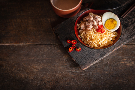 Instant noodle with pork, egg and vegetables on black bowl on the wood table there are chilli, chopstick and drinking glass placed around.の写真素材