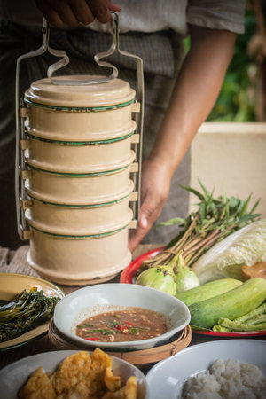 Mother holds a lunch box with shrimp paste and puts food on the table. Steamed rice, stir-fried morning glory with chili paste and omelet. Retro Thai style photo.の写真素材