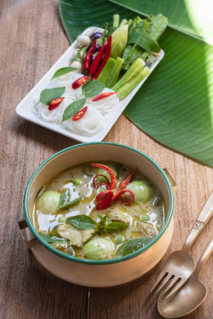 Authentic Thai green curry with tender chicken and eggplants served with rice noodles and fresh vegetables. Traditional Thai food still life on wooden table, beautifully arranged with basil leaves, red chili, and gold bowl. Perfect for Thai cuisine, local food, and Asian culture concepts.の写真素材
