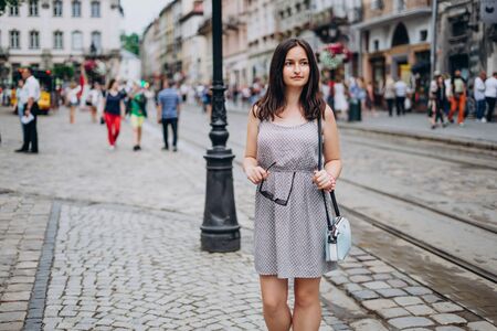 Beautiful girl in a summer dress walks the city. Brunette girl in a sundress with a handbag and glasses. Girl on the background of the city.の写真素材