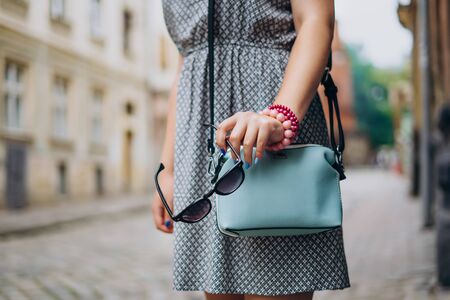 Female hand with blue handbag. Female hand with manicure and bracelets holding sunglasses. Stylish woman in dress with accessoriesの写真素材