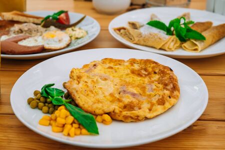 Omelet with ham, peas and corn in a cafe. Delicious omelet breakfast with cheese and peas. Omelet with vegetables on a white plate in a cozy cafeの写真素材