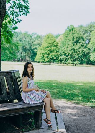 A young girl in a sundress sitting on a park bench. Beautiful woman in a summer white dress and sandals. Girl sitting on a bench on a sunny dayの写真素材