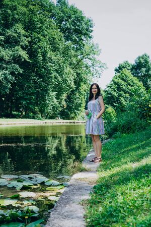 Young woman walking in the summer park. Woman in a green park near the pond. Beautiful woman in a colored sundressの写真素材