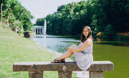 Sofia Park, Uman. Young woman sitting on a bench on the background of the lake with a fountain. The girl in a dress sits on a stone bench.の写真素材
