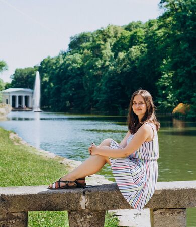 Sofia Park, Uman. Young woman sitting on a bench on the background of the lake with a fountain. The girl in a dress sits on a stone bench.の写真素材