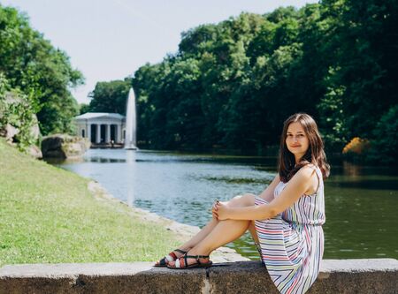 Sofia Park, Uman. Young woman sitting on a bench on the background of the lake with a fountain. The girl in a dress sits on a stone bench.の写真素材