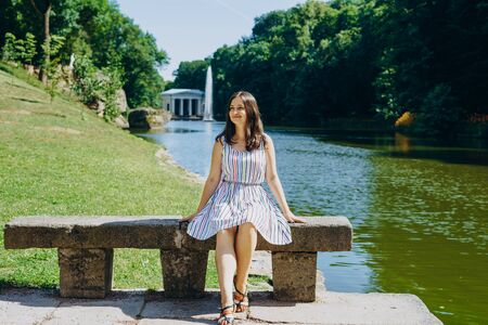 Sofia Park, Uman. Young woman sitting on a bench on the background of the lake with a fountain. The girl in a dress sits on a stone bench.の写真素材