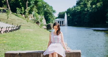 Sofia Park, Uman. Beautiful girl sitting on a park bench on the background of the lake. Girl sitting on a stone bench on the background of the river and a white gazebo.の写真素材