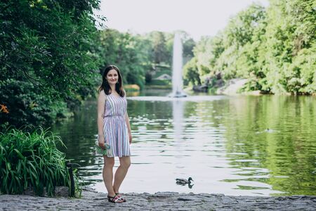 Uman, Ukraine. Smiling girl on a background of high fountain and lake. Girl in a summer dress on a background of a beautiful tall fountain. Lake in the park and splashing fountainの写真素材