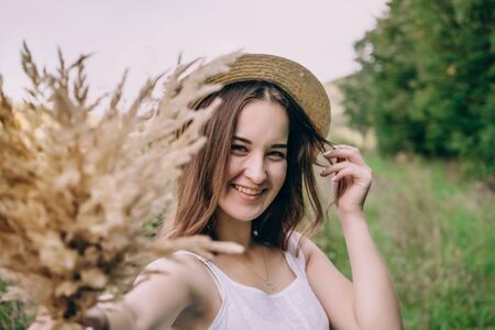 Happy young woman in a white dress in a straw hat. Smiling girl with a bouquet of dried flowers. Girl walks in a green park on a spring dayの写真素材