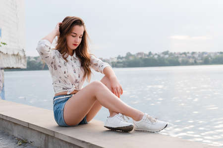 Young woman in shorts and sneakers sitting on the background of the lake. Beautiful girl posing on the background of the lake on a sunny day. Girl in casual clothesの写真素材