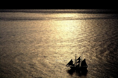 A sail boat drifts through the water off the shore of Santorini, Greece while the sun sets behind it.  の写真素材