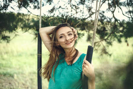 Outdoors portrait of beautiful young woman in casual green dress posing in summer garden. Smiles. On a swing.の写真素材