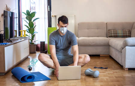 young man in mask on quarantine exercising and working at home with laptop and mobile phoneの写真素材