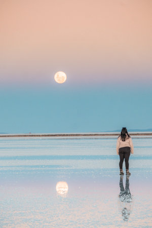 Back of a girl at Uyuni salt flats during sunset with the reflection of the moon. High quality photoの写真素材