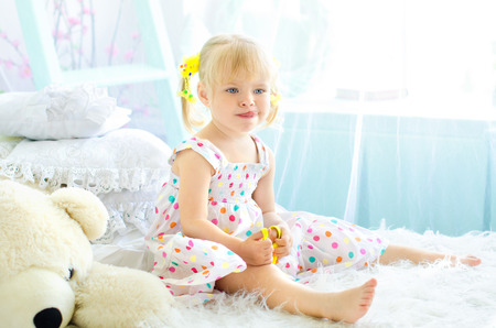 little funny girl sitting on bed in light bedroom with big white teddy bearの写真素材