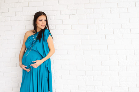 beautiful pregnant woman in a turquoise dress on white brick wall backgroundの写真素材