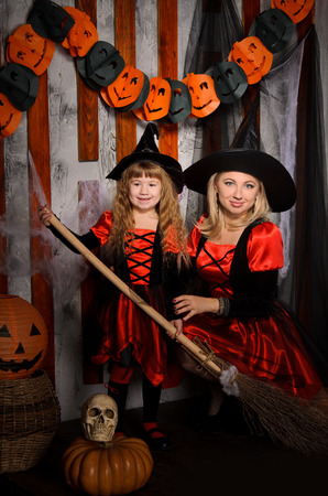 halloween scene with two attractive witches, young mother and little daughter, in costumes and hats in black and red, with broom, pumpkins and skullの写真素材