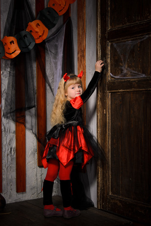 cute halloween devil girl with blonde hair with horns in costume in red and black opening old wooden door indoorsの写真素材