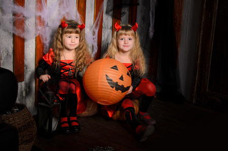 two halloween devils girls, little sisters twins, with horns in costumes in black and red with pumpkins indoorsの写真素材
