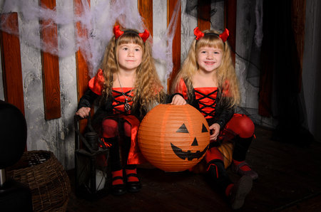 two halloween devils girls, little sisters twins, with horns in costumes in black and red with pumpkins indoorsの写真素材