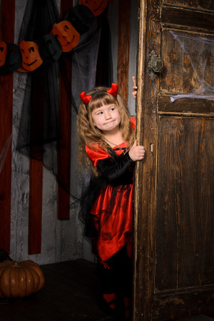 halloween devil girl in costume in black and red with horns looking out of old wooden door indoorsの写真素材