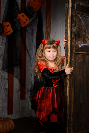 halloween devil girl in costume in red and black with horns opening old wooden door indoorsの写真素材