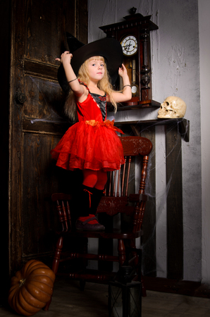 cute blonde halloween witch girl in red dress with tutu covering with large black hat in retro interior with wooden chair, clock, door and skullの写真素材