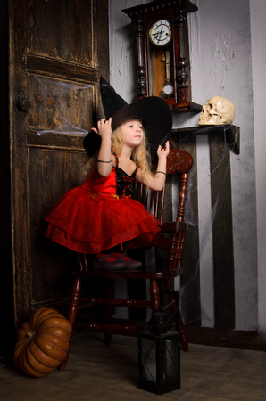 cute blonde halloween witch girl in red dress with tutu covering with large black hat in retro interior with wooden chair, clock, door and skullの写真素材