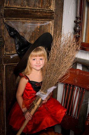 cute blonde halloween witch girl in red dress and black hat with broom at old wooden door indoorsの写真素材
