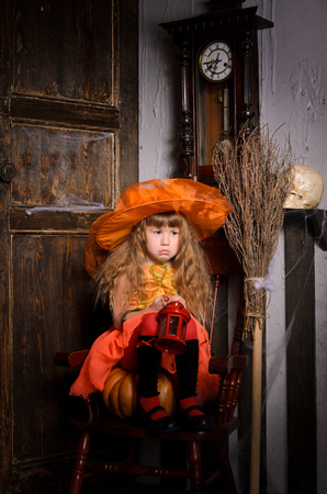 sad halloween witch girl with long hair in costume and hat in orange with lamp sitting on pumpkin on chair with broom in retro interior with old wooden door and clockの写真素材