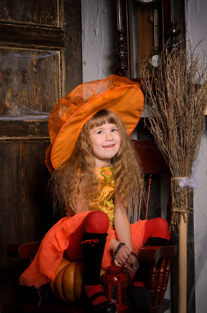 funny halloween witch girl with long hair in costume and hat in orange with lamp sitting on pumpkin on chair with broom at old wooden door indoorsの写真素材
