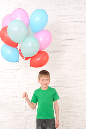 boy in green t-shirt holding a lot of colorful balloons against a white brick wallの写真素材