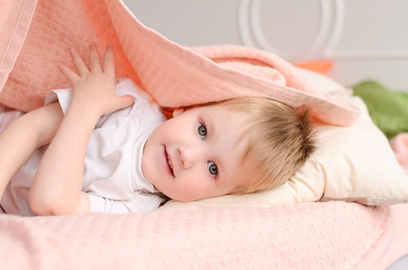 blond boy lying on a bed under a pink blanket on a white pillowの写真素材
