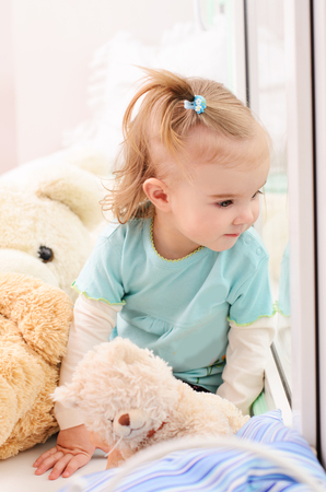little girl sitting on a window sill and looking out the window, next to stuffed toys Teddy Bearsの写真素材
